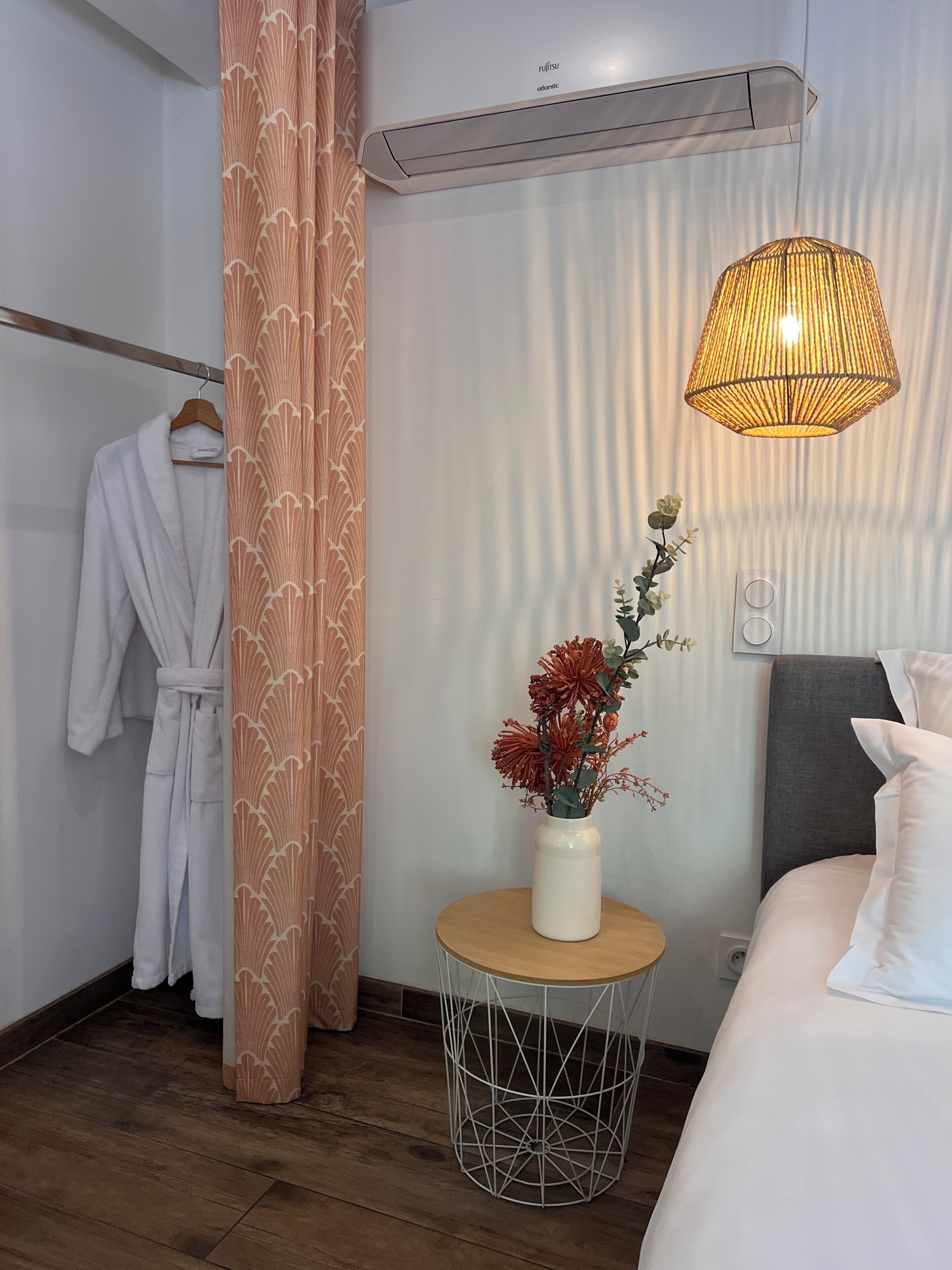 Una habitación de hotel con cama, una mesita de noche con un jarrón de flores, una bata colgada y un aire acondicionado de pared.