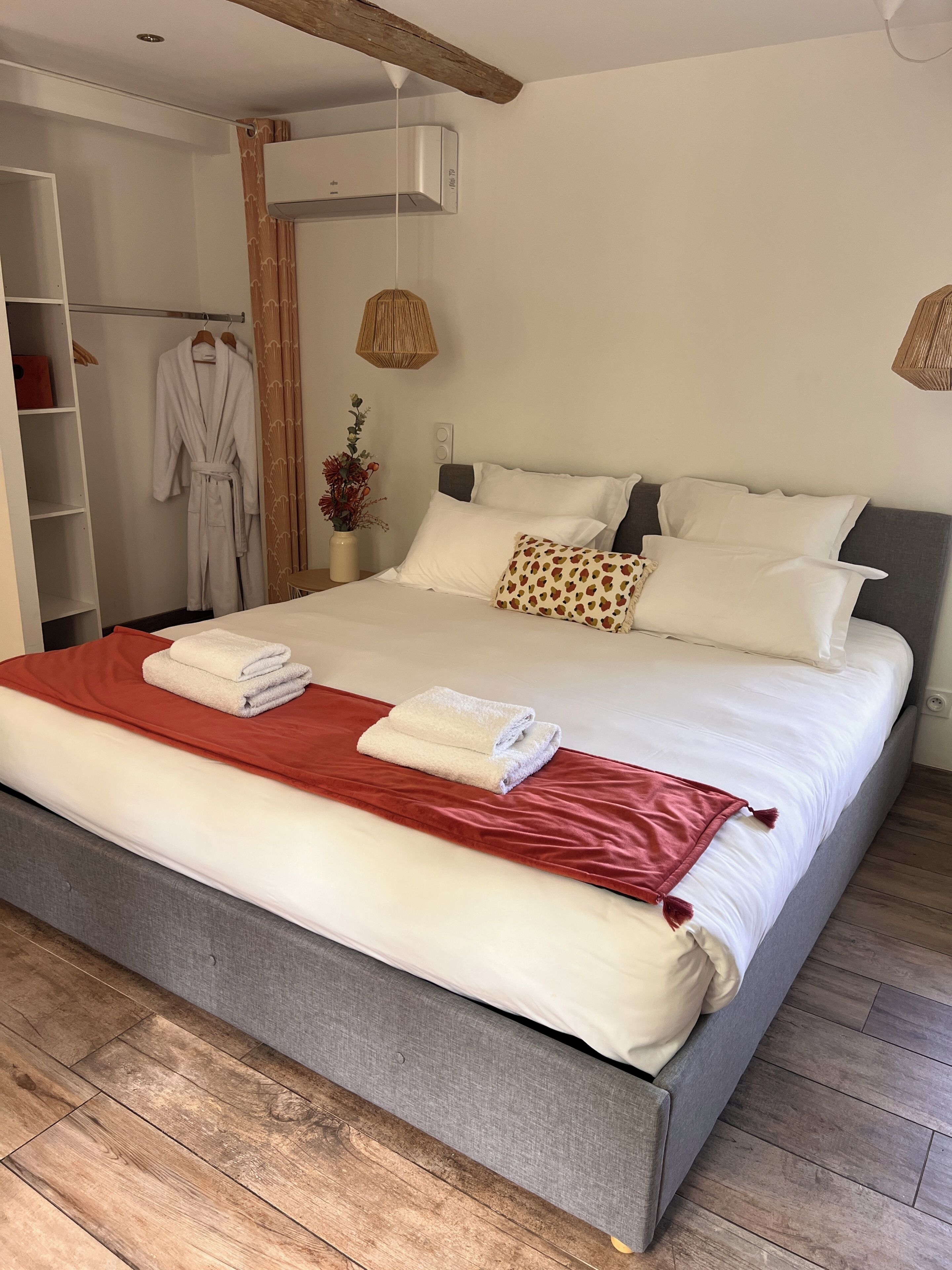 Una cama bien hecha con ropa de cama blanca y roja, un cabecero gris y suelo de madera.