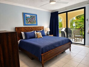 A bedroom with a wooden bed, a nightstand, a lamp, and a view of a balcony with outdoor furniture. -  DUAL Ocean Views! Beachfront Gem • Pool, Hot Tub • Steps to Sand & Dining (Christiansted)