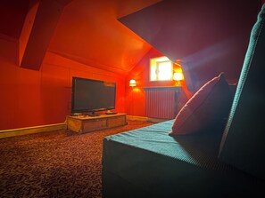 Ein gemütliches Zimmer mit einem Fernseher auf einem Holzboden, einer Couch mit Kissen und einer Lampe auf einem Tisch.