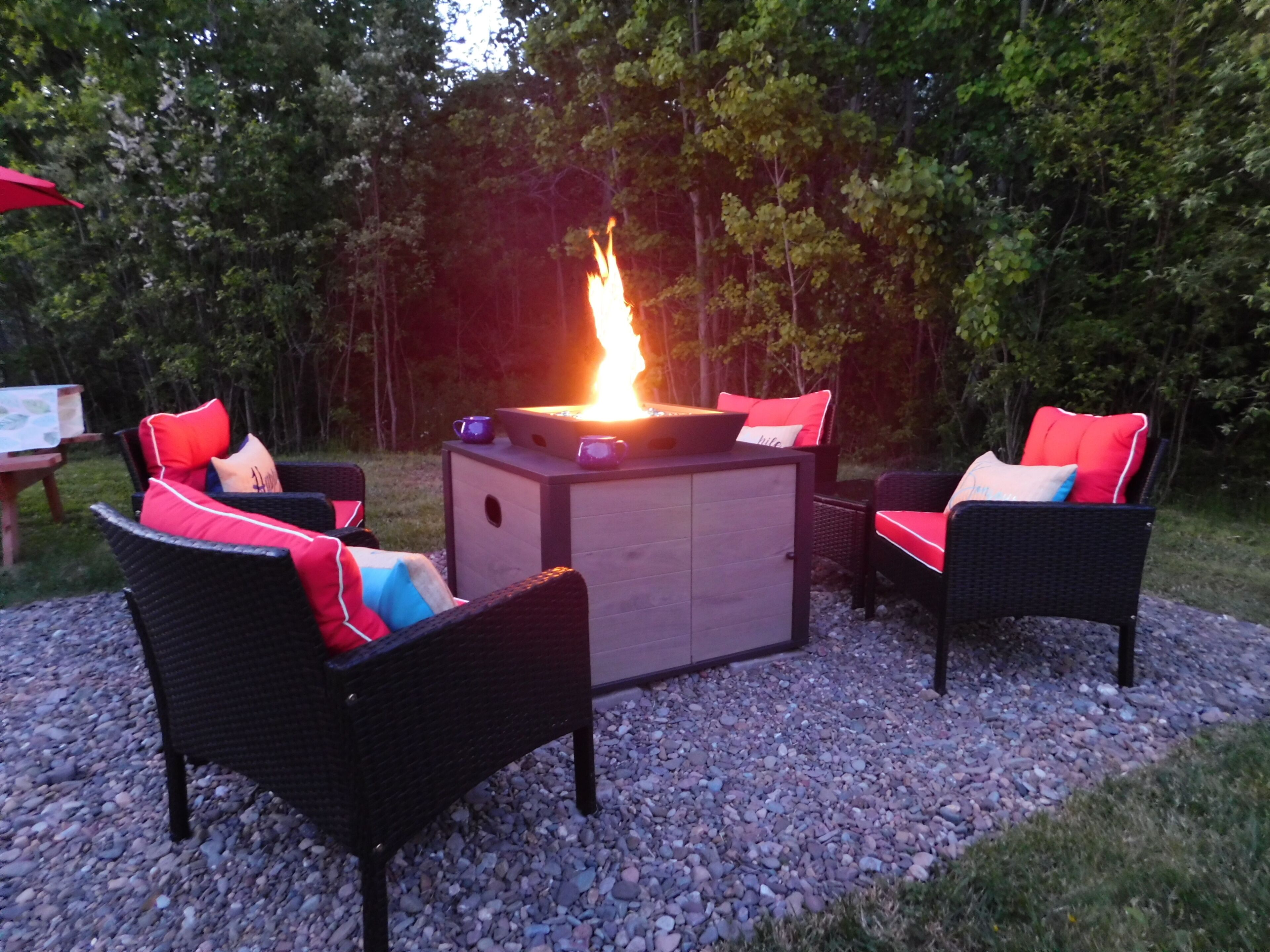 Een buitenruimte met zitplaatsen, inclusief een vuurkuil, omringd door rieten stoelen met kussens, gelegen op een grindpad met bomen op de achtergrond.