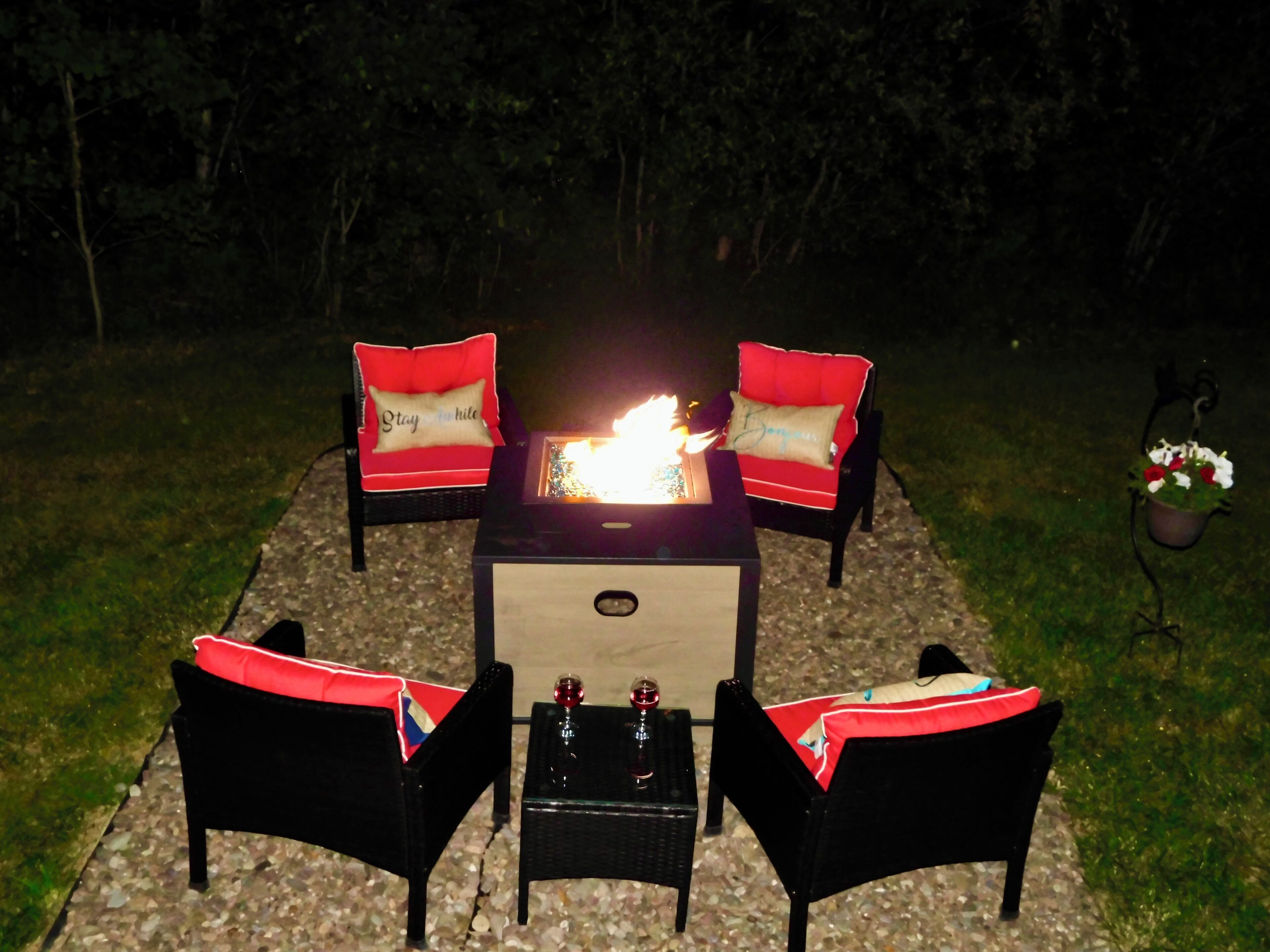Un área de estar al aire libre con una mesa de fuego, cuatro sillas y una mesita con un vaso.