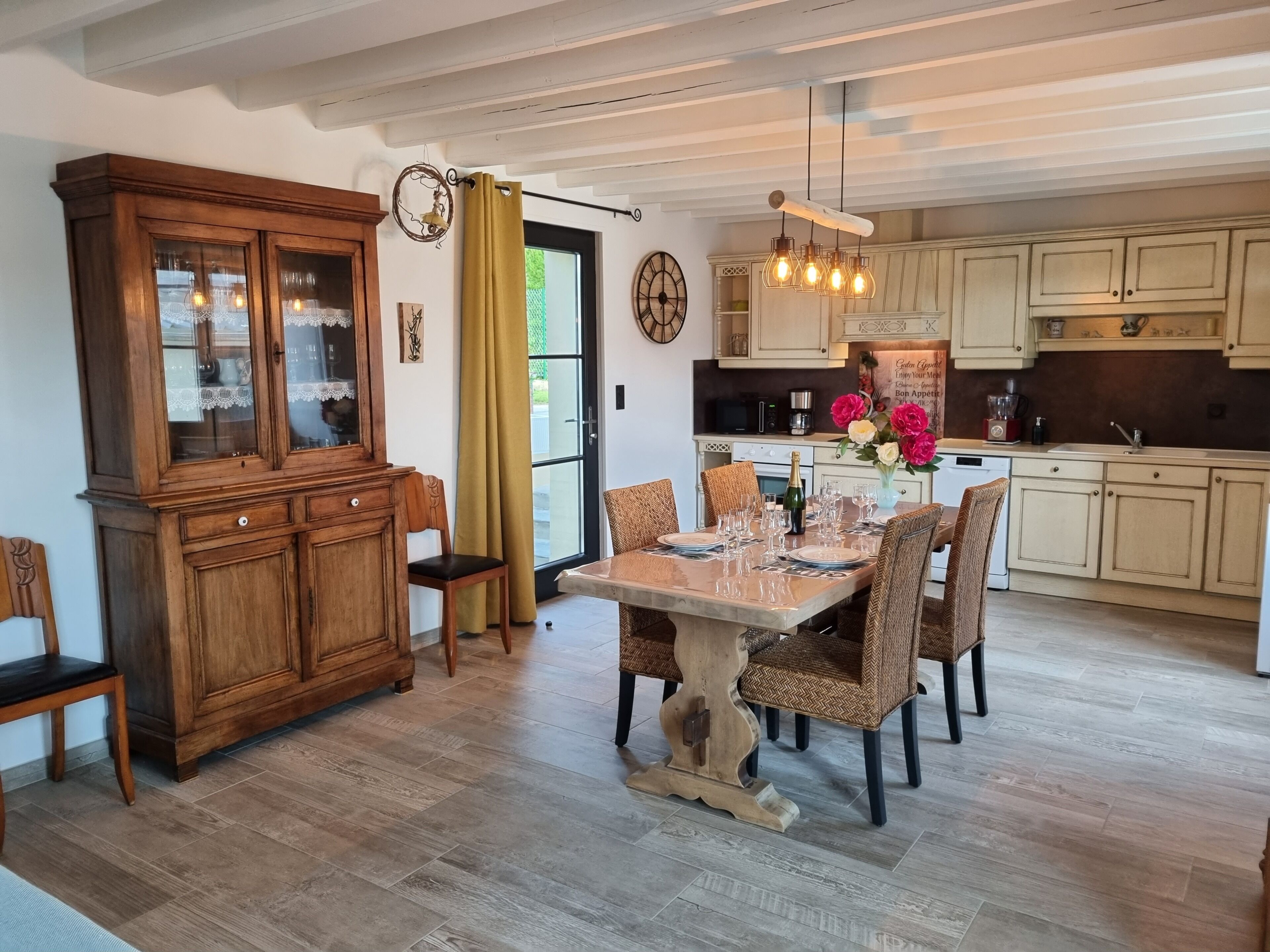 Uma sala de jantar com mesa de madeira posta para seis pessoas, cercada por cadeiras de vime, um armário de madeira e uma cozinha ao fundo.