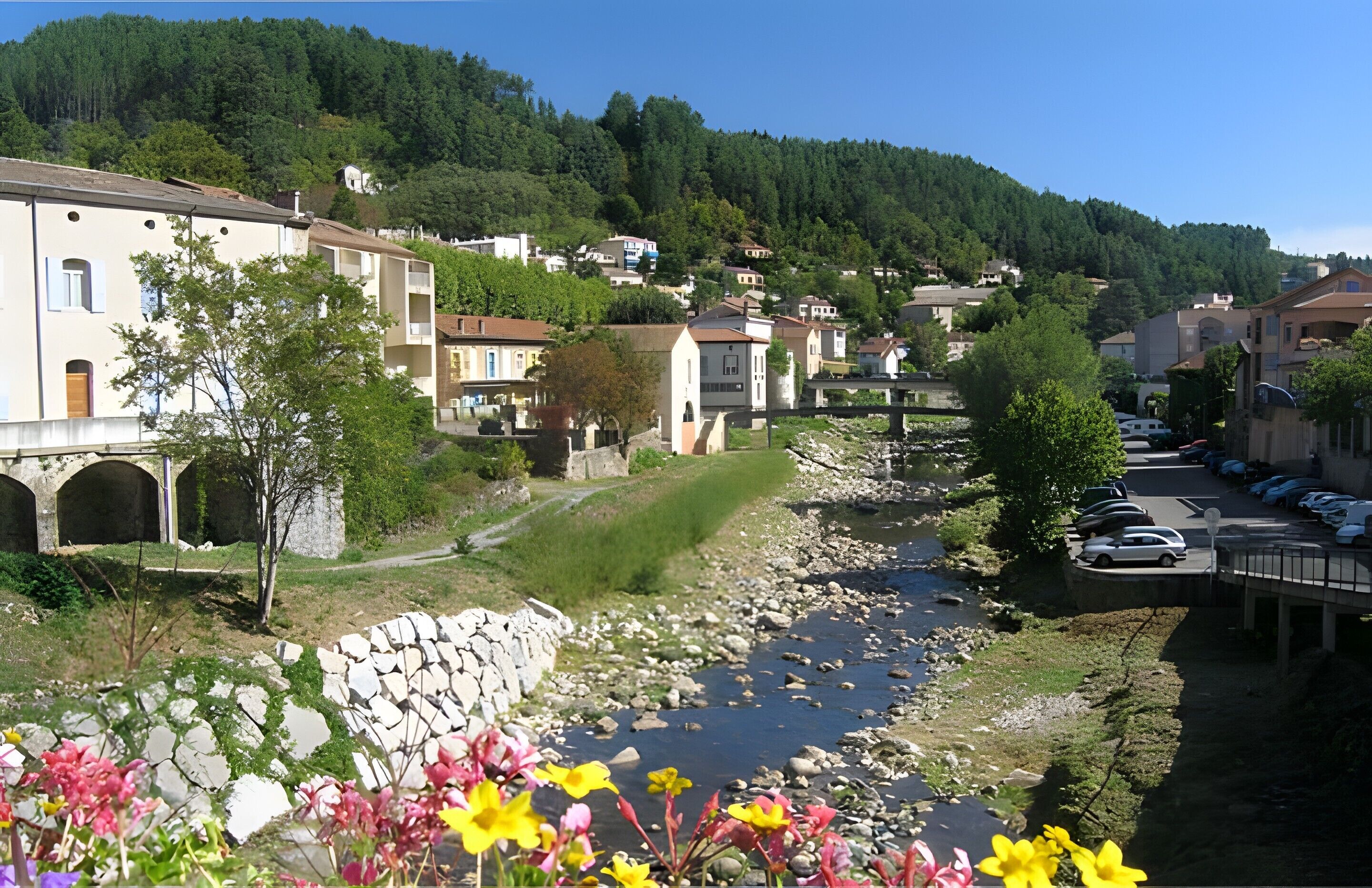 Une ville riveraine, avec des remparts en pierre, un pont et des voitures garées.