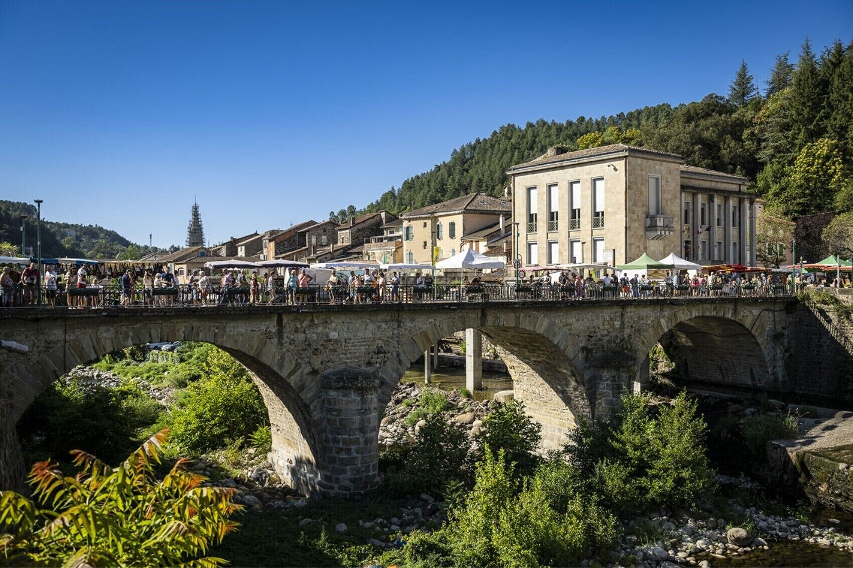 Eine Stebrücke mit Bögen über einem Fluss, umgeben von Grünflächen und Gebäuden.