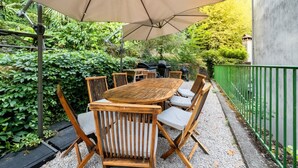 A wooden outdoor table with chairs, a large umbrella, and a barbecue grill.
