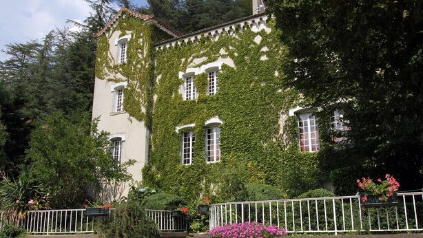 A building with a white facade partially covered in green ivy, surrounded by trees and a white fence.