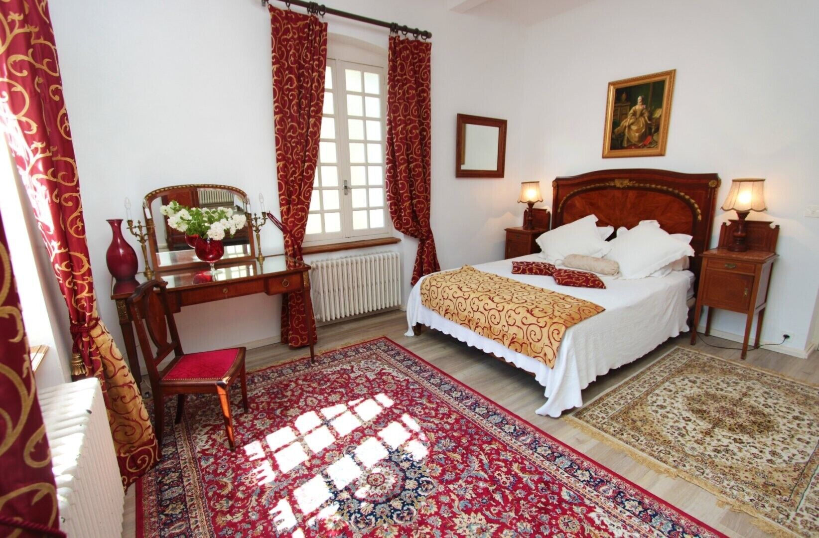 Un dormitorio con cama, escritorio, silla, radiador, ventana con cortinas, lámpara y un cuadro en la pared.