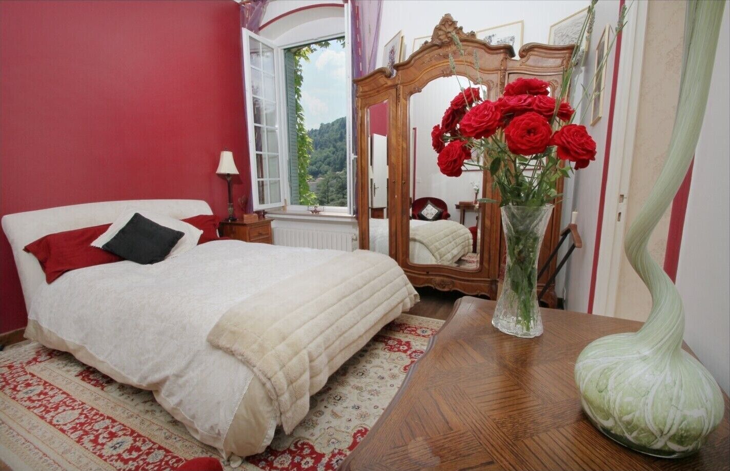 A bedroom with a bed, a mirror, a vase with flowers, and a view of greenery through a window.