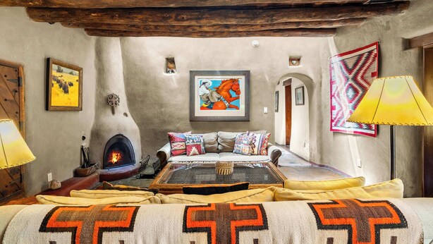 A rustic room with a fireplace, a sofa with patterned cushions, and a wall-mounted painting. - Historic Adobe, Kiva Fireplace, Walk to Canyon Rd (Santa Fe)
