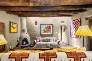 A rustic room with a fireplace, a sofa with patterned cushions, and a wall-mounted painting. - Historic Adobe, Kiva Fireplace, Walk to Canyon Rd (Santa Fe)