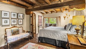 A rustic bedroom with a bed, a bench, a wooden ceiling, framed pictures on the wall, and a window with curtains. - Historic Adobe, Kiva Fireplace, Walk to Canyon Rd (Santa Fe)