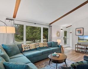 Ein modernes Wohnzimmer mit einer Couch, einem Couchtisch, einem Fernseher und Blick auf Bäume durch große Fenster.
