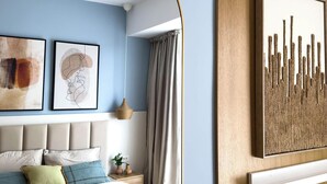 Um quarto com cama, cabeceira, uma obra de arte na parede e janela com cortinas.