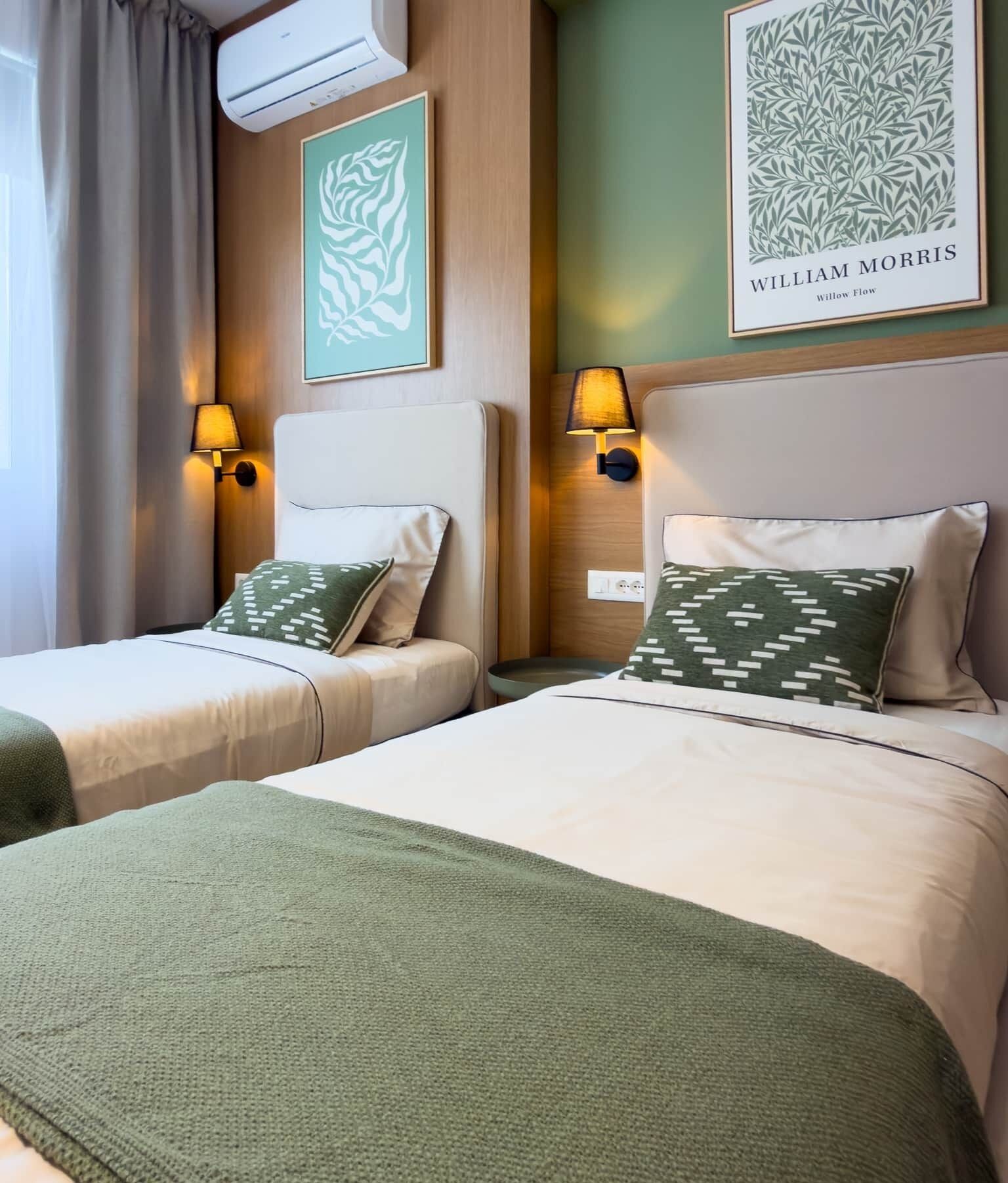 Habitación de hotel con dos camas, ropa de cama verde, una obra de arte enmarcada sobre cada cama y un aire acondicionado instalado en la pared.
