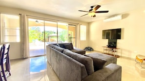 A living room with a sofa, a TV, a ceiling fan, and a sliding glass door leading to a pool area. - Spacious 5BR 4.5BA Home Private Pool Walk to Beach (Rincon)