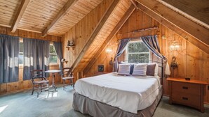 Un dormitorio con una cama grande, techo de madera, ventana con cortinas, una silla y una pequeña mesa.
