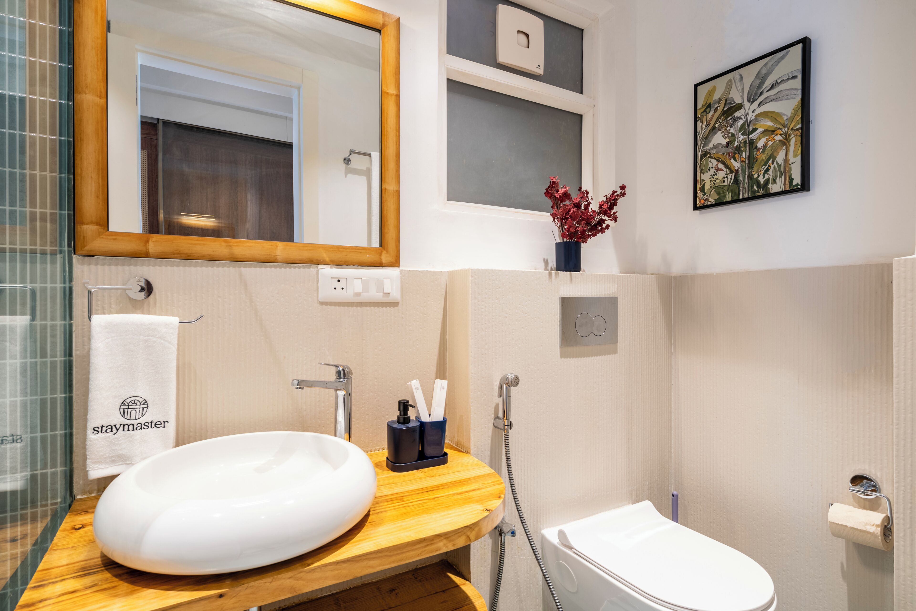 A bathroom with a wooden vanity, a white sink, a mirror, a toilet, a shower, and a framed plant.