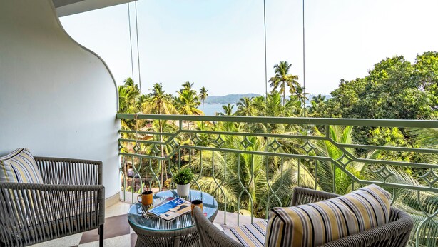 A balcony with wicker furniture, a glass table, and a view of palm trees and a distant landscape. - Staymaster Solintra 2BHK C401 (Verem)