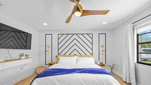 Een moderne slaapkamer met een bed, houten vloer, een plafondventilator, een tv aan de muur en een raam met gordijnen.