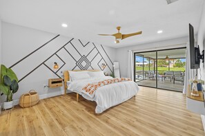 Ein Schlafzimmer mit Bett, Holzboden, Deckenventilator, Nachttisch, Lampe, Zimmerpflanze und Blick ins Freie durch eine Glastür.