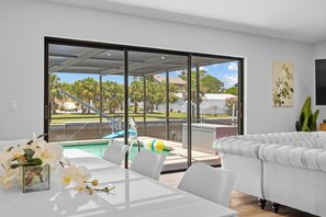 Ein modernes Wohnzimmer mit einem weißen Sofa, einem Esstisch mit Stühlen und Blick auf einen Swimmingpool und Außenbereich.