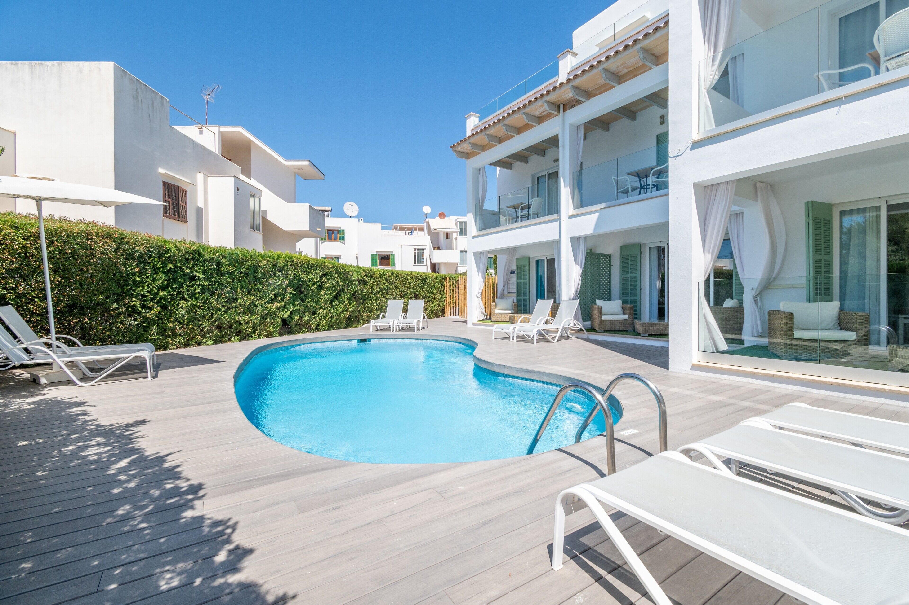 Un área de piscina moderna con sillones reclinables, una terraza de madera y un cielo azul despejado.