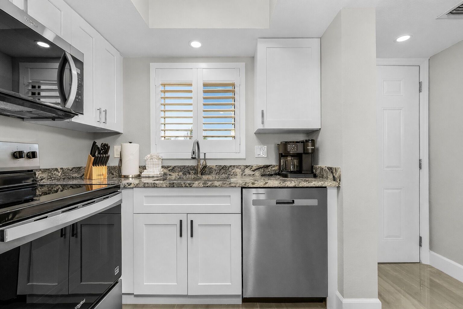 A modern kitchen with white cabinets, a stainless steel dishwasher, a built-in oven, and a window with blinds.