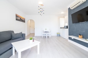 Un salon moderne avec un canapé gris, une table basse blanche, un téléviseur fixé à un mur gris et un ventilateur de plafond agrémenté de décorations suspendues.