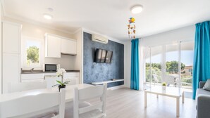 A modern living room with a dining area, a TV mounted on the wall, and a balcony with blue curtains. - 2-Bedroom Apartment 'F' with Shared Pool, Wi-Fi, and Air Conditioning (Felanitx)