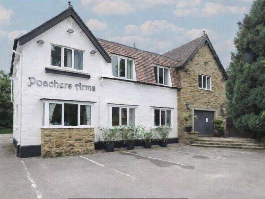 A two-story building with the name 'Poachers Arms' on the facade, a stone base, and a parking area in front.