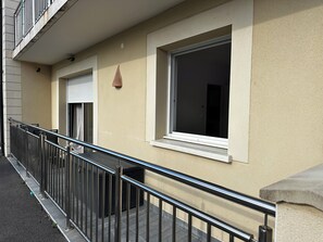A building facade with a metal balcony railing, a window with a closed shutter, and a small wall-mounted light fixture.
