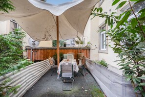 Ein überdachter Außenbereich mit Tisch und Stühlen, umgeben von Pflanzen und einem Holzzaun.