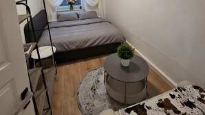 A compact bedroom with a bed, a small desk, a round table with a plant, a TV mounted on the wall, and a window with curtains.