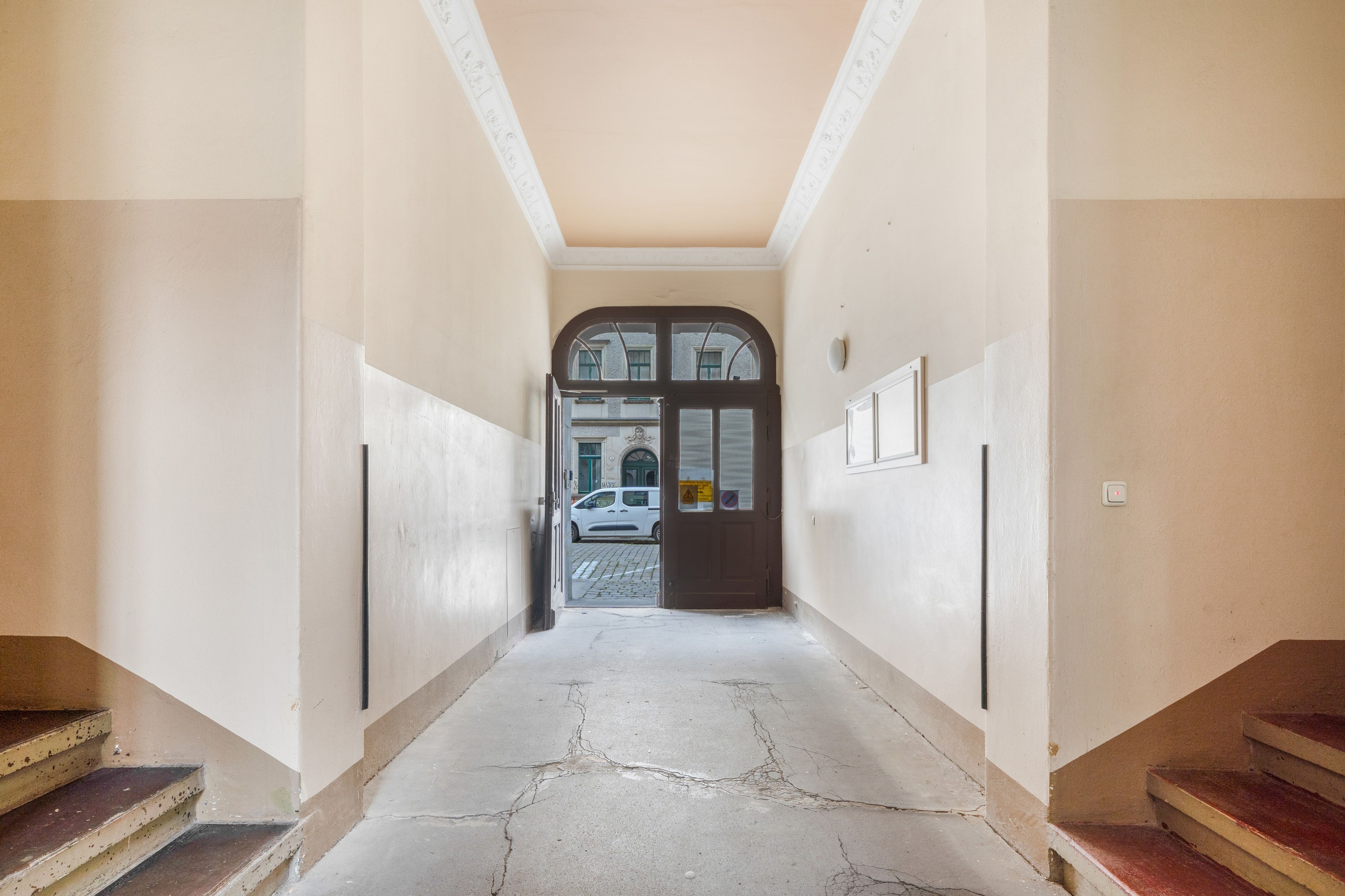 A hallway with a cracked floor, a staircase on the left, and a door at the end leading outside.