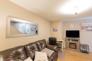 A living room with a leather couch, a flat-screen TV, a fireplace, and a large mirror on the wall. - Rosa's Sea View Serenity Stays (Stratford)
