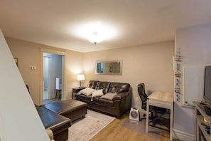 A living room with a brown leather sofa, a small table, a chair, a television, and a speaker. - Rosa's Sea View Serenity Stays (Stratford)