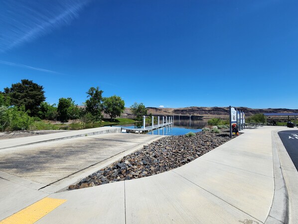 Un sendero de hormigón que lleva a un muelle sobre aguas tranquilas, con un cielo azul despejado y colinas a lo lejos.