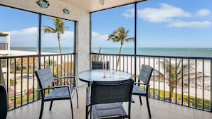 Ein Balkon mit Glastisch und Stühlen, der Blick auf einen Strand mit Palmen und das Meer freigibt.