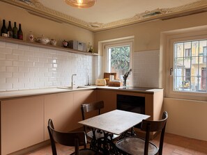 A kitchen with white subway tiles, a window with a view, a table with chairs, and a shelf with bottles. - Kiki's - A spacious house, with a garden, in the Langhe. (Castagnole delle Lanze)