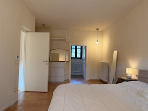 Um quarto com cama, guarda-roupa, espelho, radiador e porta que dá para outro espaço.