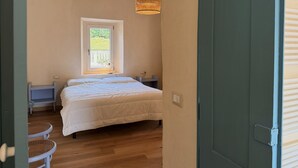 Un dormitorio con cama, ventana, mesita de noche, lámpara y una silla de mimbre.