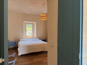 A bedroom with a bed, a window, a bedside table, a lamp, and a wicker chair. - Kiki's - A spacious house, with a garden, in the Langhe. (Castagnole delle Lanze)