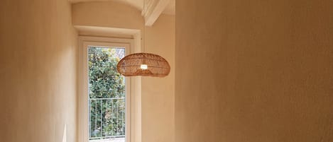Een smalle gang met een raam, een geweven lamp en een witte deur.
