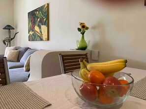 Ein Essbereich mit Tisch, Stühlen, einer Obstschale, einer Blumenvase und einem Bild an der Wand.