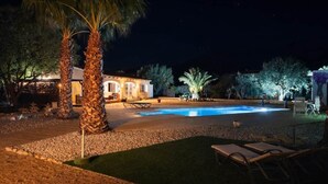 Une vue nocturne d’une maison avec une piscine, des palmiers et un espace salon extérieur.