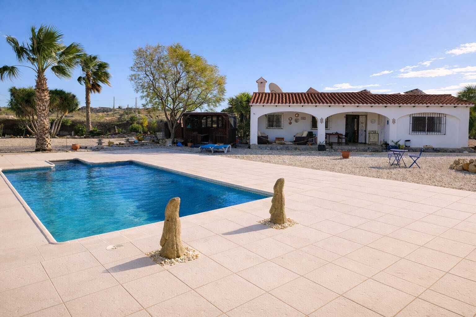 Une piscine bordée de deux bornes en pierre, un bâtiment blanc avec un toit en tuiles, et des palmiers en arrière-plan.