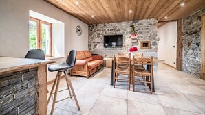 Un interior acogedor con pared de piedra, techo de madera, televisor montado, mesa de comedor con sillas y un sofá.