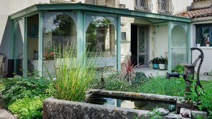 Une véranda avec une structure en verre, un petit étang et une végétation luxuriante tout autour.