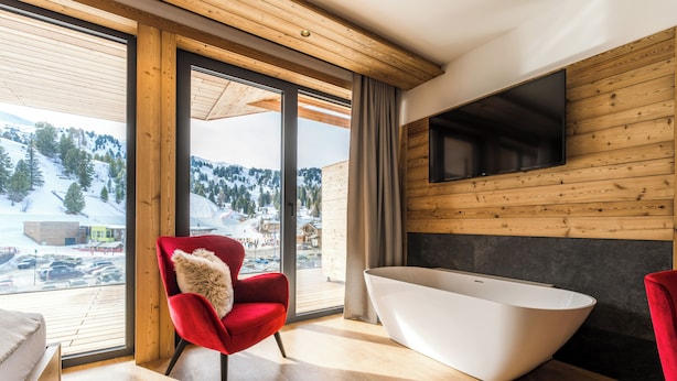 Une salle de bain moderne avec une baignoire sur pieds, un fauteuil rouge agrémenté d’un plaid en fourrure et une grande fenêtre donnant sur un paysage enneigé.